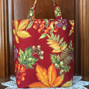 May include: A red tote bag with a fall-themed pattern featuring leaves, berries, and acorns. The bag has matching handles and is made of fabric. The design includes shades of red, orange, yellow, and green.