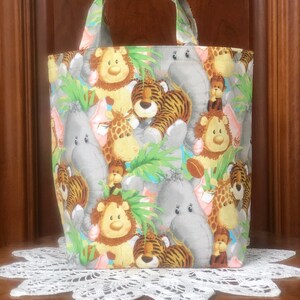 May include: A fabric tote bag featuring a repeating pattern of cartoon jungle animals including gray elephants, orange and black striped tigers, yellow lions, and brown monkeys, accented with green palm leaves and two matching fabric handles.