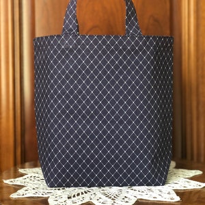 May include: A navy blue tote bag with a white diamond pattern. The bag has two handles and is sitting on a white lace doily. The background is a wooden door.