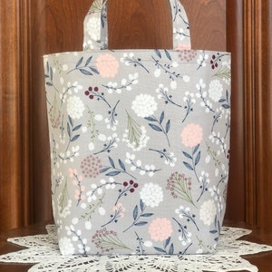 May include: A gray tote bag with a floral print featuring white, pink, and blue flowers and dark red berries on a light gray background.