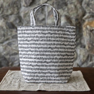 Puede incluir: Un bolso tote blanco cubierto de notaci&oacute;n musical negra. El bolso tiene dos asas y est&aacute; colocado sobre un tapete beige. Est&aacute; sobre una mesa de madera marr&oacute;n oscuro, con una pared de piedra al fondo.