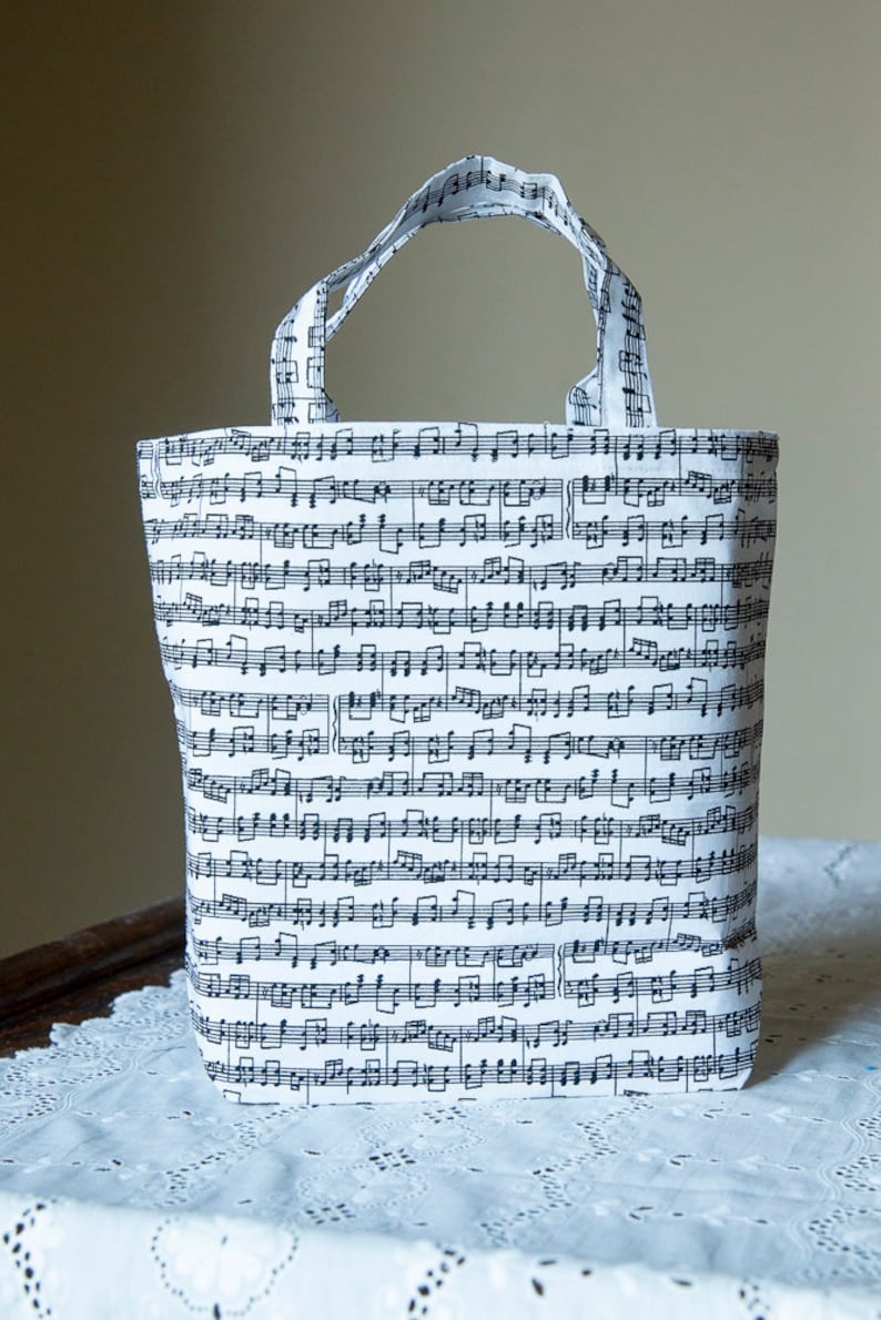 Puede incluir: Un bolso tote blanco con un patr&oacute;n de partituras musicales negras. El bolso tiene dos asas y est&aacute; hecho de tela. Las notas musicales est&aacute;n impresas en todo el bolso, creando un dise&ntilde;o &uacute;nico y art&iacute;stico. El bolso est&aacute; sobre un mantel de encaje blanco.