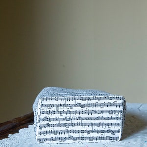 Puede incluir: Una caja rectangular cubierta de tela blanca estampada con notas musicales negras. La caja est&aacute; sobre un mantel de encaje blanco. El fondo es de color neutro.