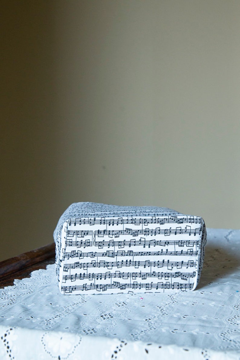 Puede incluir: Una caja rectangular cubierta de tela blanca estampada con notas musicales negras. La caja est&aacute; sobre un mantel de encaje blanco. El fondo es de color neutro.
