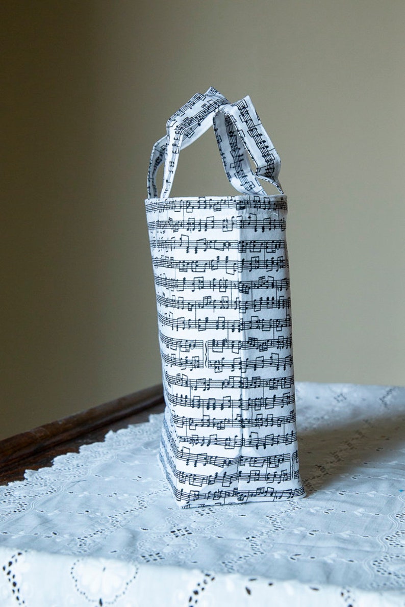 Puede incluir: Un bolso tote alto con asas, cubierto con un patr&oacute;n de partituras musicales en blanco y negro. El bolso es de tela y se apoya sobre un mantel de encaje blanco. Las asas tambi&eacute;n est&aacute;n hechas de la misma tela de partituras musicales.