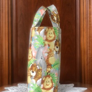 May include: A fabric tote bag featuring a repeating pattern of cartoon jungle animals including lions, tigers, giraffes, and elephants amidst green foliage on a light gray background, with two matching fabric handles at the top of the bag.
