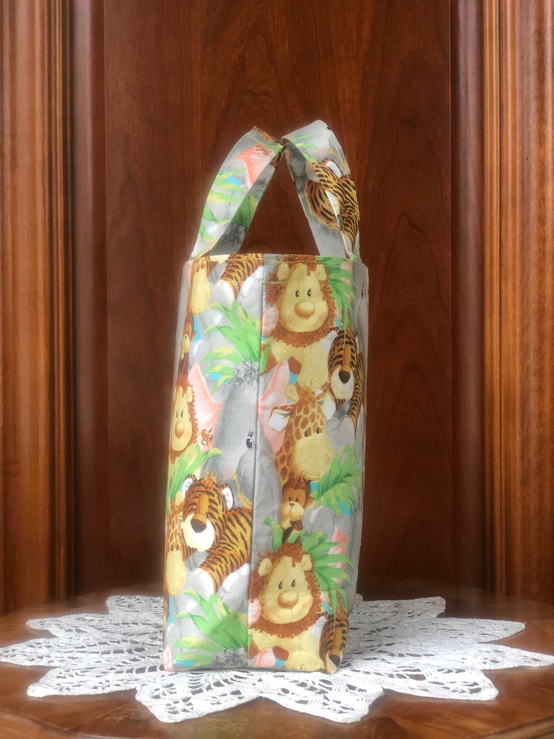 May include: A fabric tote bag featuring a repeating pattern of cartoon jungle animals including lions, tigers, giraffes, and elephants amidst green foliage on a light gray background, with two matching fabric handles at the top of the bag.