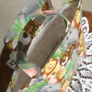 May include: A fabric tote bag featuring a colorful jungle animal print with tigers, lions, giraffes, and hippos on a light gray background. The bag has two matching fabric handles and a plain white interior lining, showing an open top design.