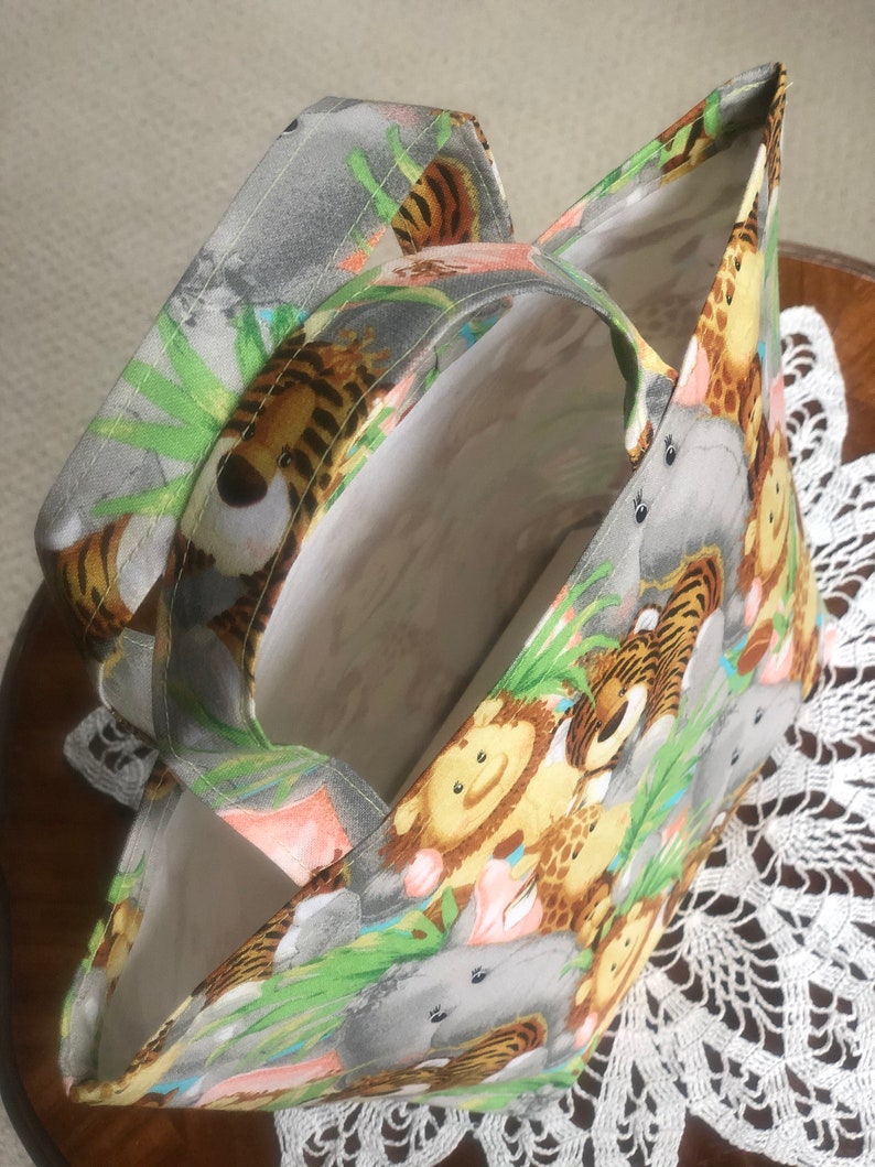 May include: A fabric tote bag featuring a colorful jungle animal print with tigers, lions, giraffes, and hippos on a light gray background. The bag has two matching fabric handles and a plain white interior lining, showing an open top design.