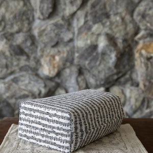 Puede incluir: Un coj&iacute;n rectangular cubierto con tela estampada con notas musicales negras sobre un fondo blanco. El coj&iacute;n descansa sobre un mantel individual color crema, sobre una mesa de madera oscura. El fondo es una pared de piedra.