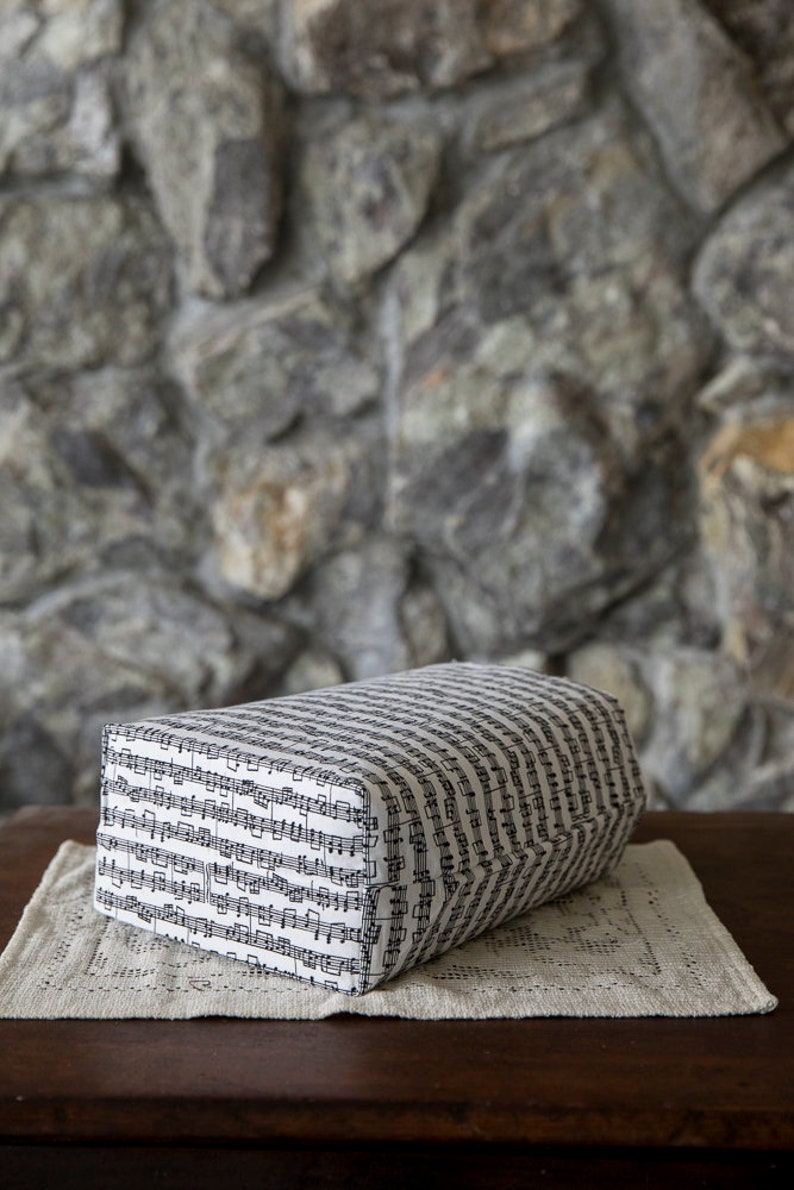 Puede incluir: Un coj&iacute;n rectangular cubierto con tela estampada con notas musicales negras sobre un fondo blanco. El coj&iacute;n descansa sobre un mantel individual color crema, sobre una mesa de madera oscura. El fondo es una pared de piedra.
