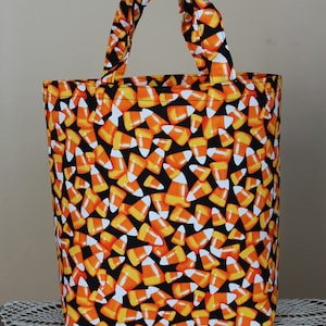 May include: A fabric tote bag with a repeating pattern of candy corn on a black background. The bag has a rectangular shape with a matching candy corn patterned handle. The bag is designed for carrying items and has a Halloween theme.