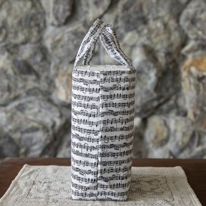 Puede incluir: Un bolso tote cil&iacute;ndrico con un patr&oacute;n de notas musicales en blanco y negro. El bolso tiene una base rectangular y dos asas. Est&aacute; sobre un mantel individual crema, similar al encaje, sobre una mesa de madera oscura.