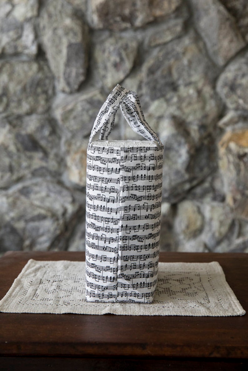 Puede incluir: Un bolso tote cil&iacute;ndrico con un patr&oacute;n de notas musicales en blanco y negro. El bolso tiene una base rectangular y dos asas. Est&aacute; sobre un mantel individual crema, similar al encaje, sobre una mesa de madera oscura.