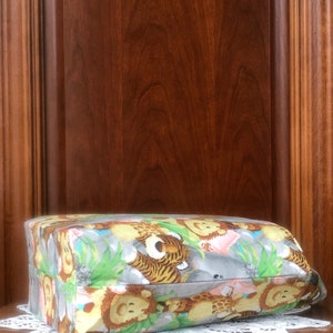 May include: A rectangular fabric pouch featuring a repeating pattern of cartoon jungle animals including lions, tigers, and giraffes set against a gray background with green foliage accents. The pouch has a structured shape and a side carry handle.