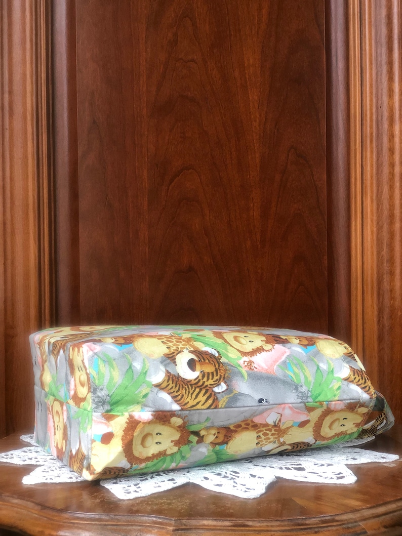 May include: A rectangular fabric pouch featuring a repeating pattern of cartoon jungle animals including lions, tigers, and giraffes set against a gray background with green foliage accents. The pouch has a structured shape and a side carry handle.