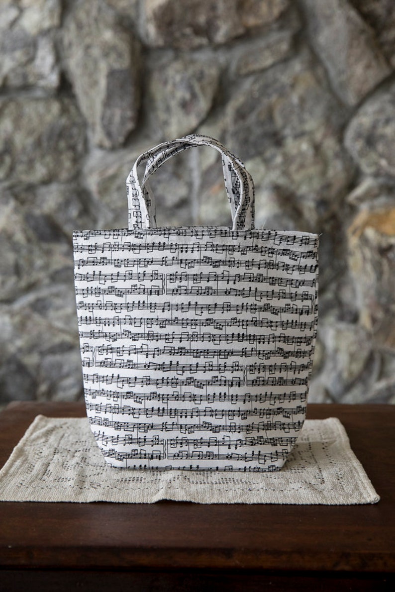Puede incluir: Un bolso tote blanco cubierto de notaci&oacute;n musical negra. El bolso tiene dos asas y est&aacute; colocado sobre un tapete beige. Est&aacute; sobre una mesa de madera marr&oacute;n oscuro, con una pared de piedra al fondo.