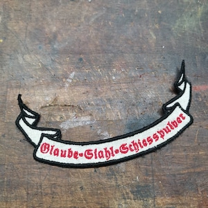 Könnte beinhalten: Weißer gestickter Aufnäher mit schwarzem Umriss und roter Beschriftung. Der Aufnäher lautet "Glaube-Glahl-Schliesspulver".