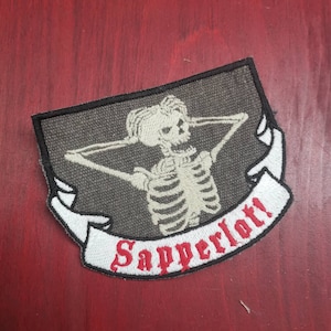 May include: A black and white embroidered patch with a skeleton holding its head in its hands. The skeleton is on a gray background with a white banner that says "Sapperlot!" in red lettering.