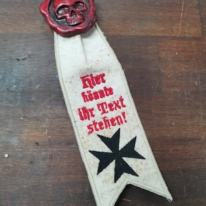 May include: A white fabric bookmark with red embroidery that reads "Hier könnte Ihr Text stehen!" and a black cross. The bookmark has a red wax seal with a skull on top.