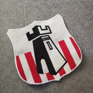 May include: A shield-shaped patch with a white background and a black tower design. The lower section has alternating red and white vertical stripes. The patch is likely for decorative purposes or to be sewn onto clothing.