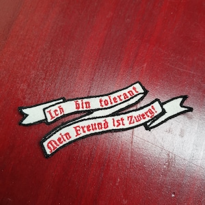 Könnte beinhalten: Zwei weiße Stoffbanner mit schwarzen Rändern und rotem Sticktext. Das obere Banner lautet "Ich bin tolerant". Das untere Banner lautet "Mein Freund ist Zwerg!"