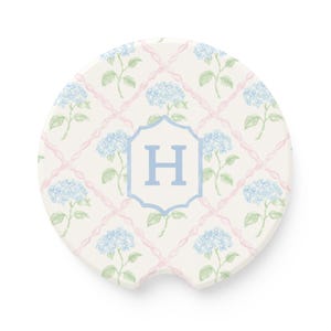 May include: A white car coaster with a floral design featuring blue hydrangeas and pink ribbon lattice. The center has a light blue shield with the letter "H".