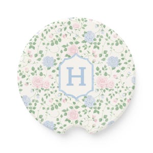 Può includere: Sottobicchiere rotondo per auto con un motivo floreale di rose rosa e ortensie blu su sfondo crema. Il sottobicchiere presenta la lettera "H" azzurra all'interno di uno scudo bianco e azzurro.