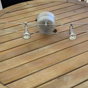 May include: A silver track lighting fixture with four adjustable spotlights and a central cylindrical component. The fixture is mounted on a wooden table with a natural finish. The spotlights are angled, suggesting they can be adjusted to illuminate different areas.