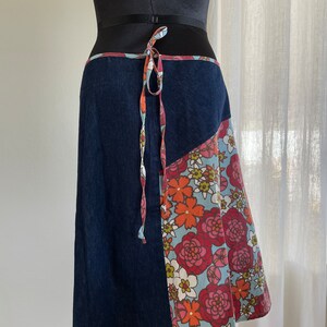 K&ouml;nnte beinhalten: Ein Wickelrock aus Jeans mit einem floralen Print-Panel. Der Rock hat einen Bindeverschluss an der Taille und eine dunkelblaue Jeansbasis.