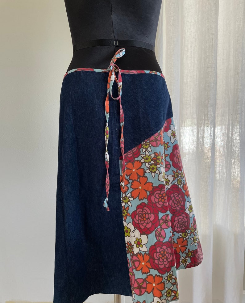 K&ouml;nnte beinhalten: Ein Wickelrock aus Jeans mit einem floralen Print-Panel. Der Rock hat einen Bindeverschluss an der Taille und eine dunkelblaue Jeansbasis.