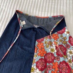 K&ouml;nnte beinhalten: Ein Jeansrock mit einem floralen Print-Panel. Der Rock hat einen Wickelstil und einen Bindeverschluss. Der florale Print ist in Rot-, Orange-, Rosa- und Gr&uuml;nt&ouml;nen auf einem hellblauen Hintergrund.