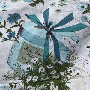 May include: A vintage fabric with a blue and white floral pattern. A blue gift box with a blue and white ribbon is in the center of the fabric. The box has a label that says "Mme. Ernest Millier, New York".