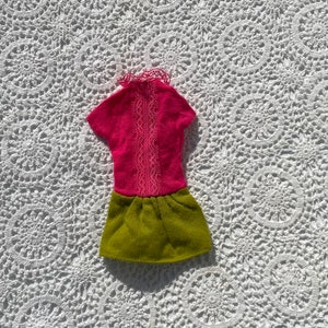 May include: A pink and green doll dress with a lace trim. The dress has a short sleeve top and a gathered skirt.