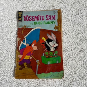 May include: A vintage comic book cover featuring Yosemite Sam and Bugs Bunny. The cover is yellow, orange, and red with the title "Yosemite Sam and Bugs Bunny" in black text. The comic book is from the Gold Key Comics series.