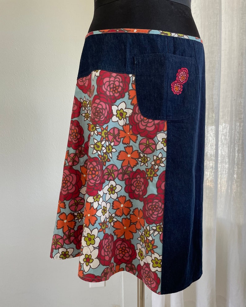 K&ouml;nnte beinhalten: Ein Jeansrock mit einem floralen Print-Panel und einer einzigen Tasche auf der rechten Seite. Die Tasche hat ein kleines gesticktes Blumenmuster.