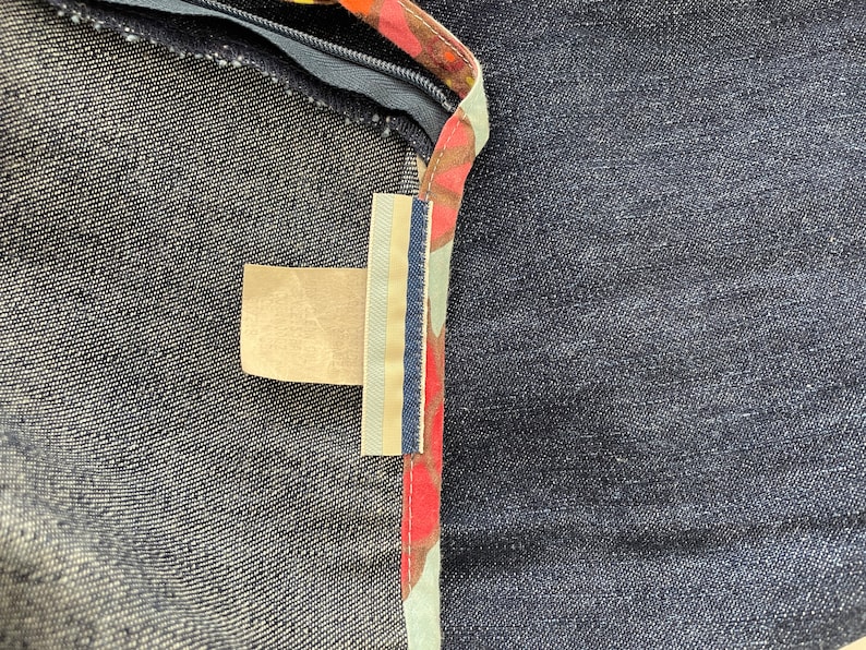 K&ouml;nnte beinhalten: Nahaufnahme eines Jeansstoffes mit einem braunen, roten und wei&szlig;en Musterbesatz. Ein kleines, rechteckiges, beigefarbenes Etikett ist am Besatz befestigt.