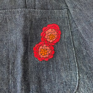 K&ouml;nnte beinhalten: Nahaufnahme einer Jeans-Tasche mit zwei gestickten roten Blumen. Die Blumen haben rosa und orangefarbene Akzente.