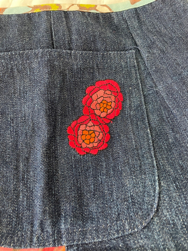 K&ouml;nnte beinhalten: Nahaufnahme einer Jeans-Tasche mit zwei gestickten roten Blumen. Die Blumen haben rosa und orangefarbene Akzente.