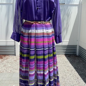 May include: A purple long-sleeved shirt with a collar and buttons, paired with a multi-colored striped skirt. The skirt has a gold belt.