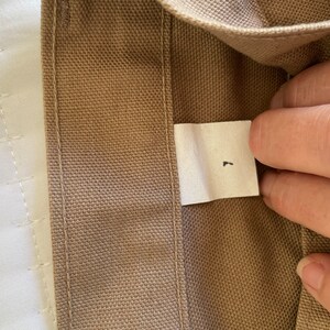 Op de afbeelding: Een close-up van een bruine canvas stof met een wit label waarop "L" staat.