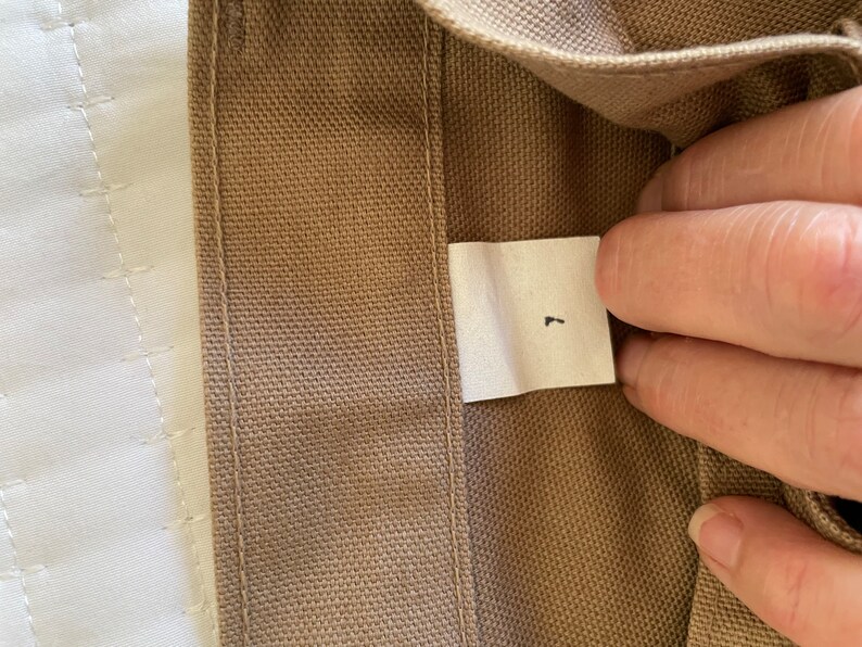 Op de afbeelding: Een close-up van een bruine canvas stof met een wit label waarop "L" staat.