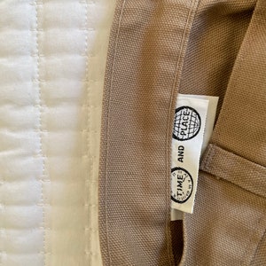 Op de afbeelding: Een beige canvas draagtas met een wit label waarop "TIME AND PLACE" staat met een wereldbolpictogram.