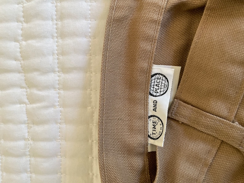 Op de afbeelding: Een beige canvas draagtas met een wit label waarop "TIME AND PLACE" staat met een wereldbolpictogram.
