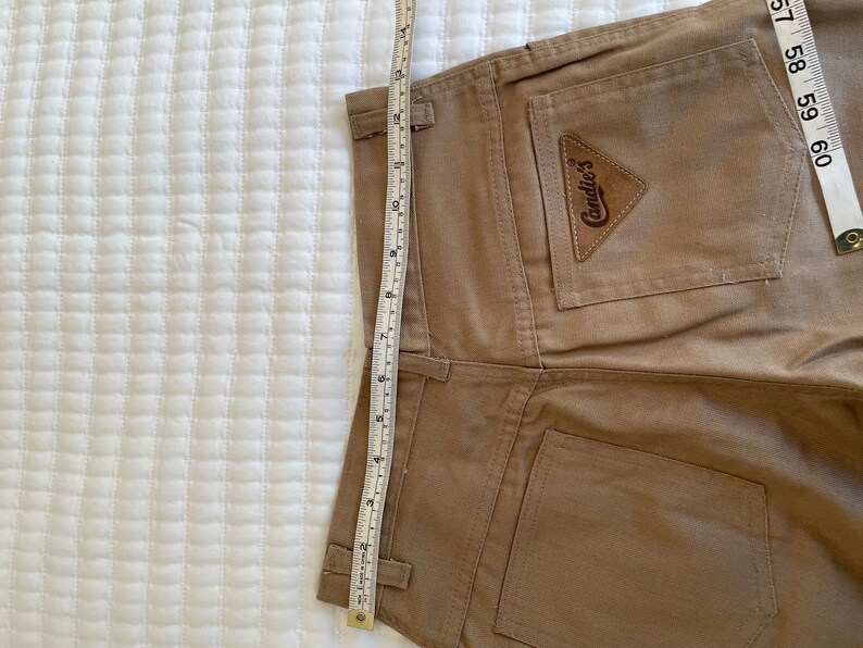 Op de afbeelding: Een bruine corduroy broek met een achterzak die een bruin leren patch heeft met de tekst "Candie's" in witte letters.