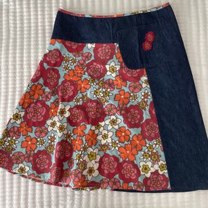 K&ouml;nnte beinhalten: Ein Rock aus blauem Denim und mit Blumenmuster, der an der Seite eine gro&szlig;e Tasche hat. Der Rock hat einen blauen Denim-Bund und ein Blumenmuster, das rote, orange, wei&szlig;e und gr&uuml;ne Blumen enth&auml;lt.
