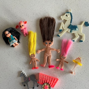 May include: A collection of vintage troll dolls, including a pink troll with a pig nose, a yellow troll with long hair, a brown troll with long hair, a pink troll with long hair, and a blue troll with long hair. The dolls are arranged on a white surface with a white horse, a gray horse and cart, and a birthday cake.