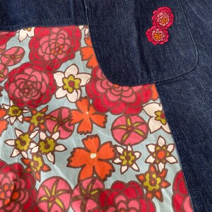 K&ouml;nnte beinhalten: Ein blauer Jeansrock mit einem floralen Print-Panel und einer einzigen Ges&auml;&szlig;tasche. Die Tasche hat zwei gestickte rosa Blumen.