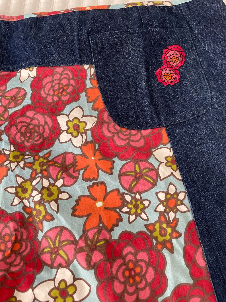 K&ouml;nnte beinhalten: Ein blauer Jeansrock mit einem floralen Print-Panel und einer einzigen Ges&auml;&szlig;tasche. Die Tasche hat zwei gestickte rosa Blumen.