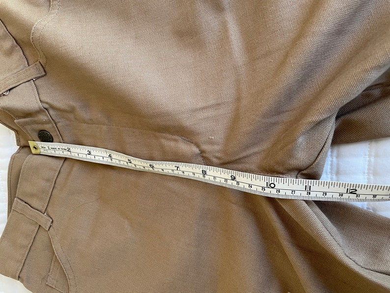 Op de afbeelding: Een bruine broek met een meetlint over de tailleband. Het lint geeft 12 inch aan.